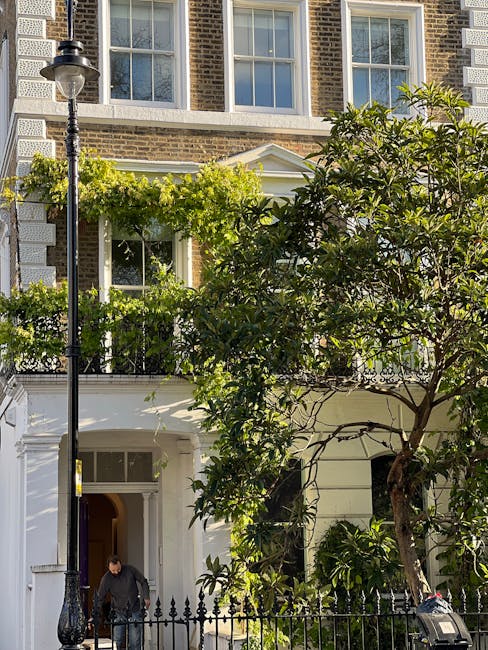 A narrow residential street featuring a row of terraced brick houses with white-framed windows and small front gardens filled with a variety of potted plants, including pink hydrangeas, green shrubs, 