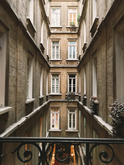 A narrow courtyard garden situated between tall, historic stone buildings with multiple white-framed sash windows. The garden features a reflective water feature or pool along the base of the building